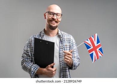 Young caucasian bald beard man in glasses holds an United Kingdom flag and clipboard with happy facial expression isolated on gray background  - Shutterstock ID 2128926749