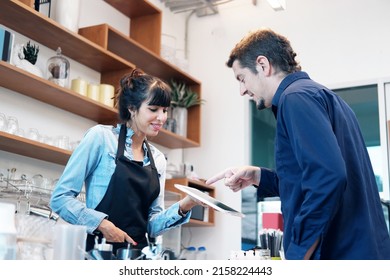 Young Caucasian barista woman is holding tablet for customers to choose a drink menu on tablet and payment at counter bar at coffee shop. Technology of digital for start up business concept.