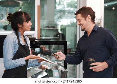 Young Caucasian barista woman is holding tablet for customer using smart phone scan QR code on tablet for payment at counter bar at coffee shop. Technology of digital pay without money concept - Powered by Shutterstock