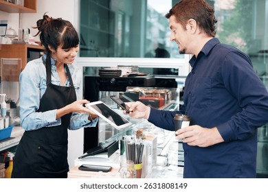 Young Caucasian barista woman holding tablet for customer using smart phone scan QR code for payment at counter bar at coffee shop. Technology of digital pay without money with mobile banking