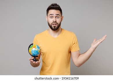 Young caucasian bearded geography student traveler indignant confused man in yellow basic t-shirt hold in palms Earth world globe spread hands look camera isolated on grey background studio portrait