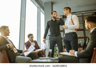 young caucasian bearded man in black formal shirt and man in white shirt stand giving speech to co-workers, holding paper document in hands. isolated in modern office with panoramic window - Shutterstock ID 1628518831