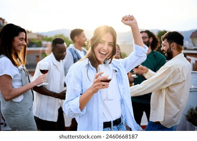 Young Caucasian beautiful woman posing holding wine with friends dancing at rooftop. Excited people have fun outdoor in dusk. Lively pretty female looking smiling at camera celebrating summer party - Powered by Shutterstock