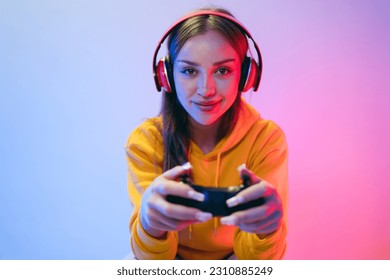 A young Caucasian beautiful woman in a yellow hoodie playing a video game using a joystick and headphones with fun, focus, surprise, and a winning victory face with blue and red neon light. - Powered by Shutterstock