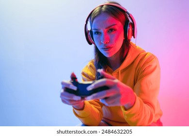 A young Caucasian beautiful woman in a yellow hoodie playing a video game using a joystick and headphones with fun, focus, surprise, and a winning victory face with blue and red neon light. - Powered by Shutterstock