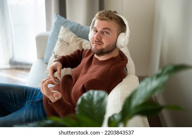 Young caucasian blond curly hair man listening to music with big white handsfree headphones sitting on sofa at home on neutral white background. Handsome adult guy portrait lifestyle.Copyspace.