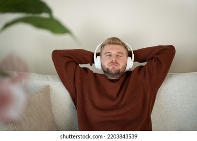 Young caucasian blond curly hair man listening to music with big white handsfree headphones sitting on sofa at home on neutral white background. Handsome adult guy portrait lifestyle.Copyspace.