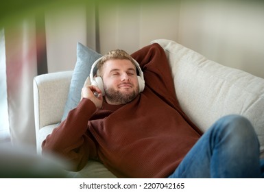 Young caucasian blond curly hair man enjoy listening to music with big white handsfree headphones lying on sofa at home. Handsome adult guy in modern interior,lifestyle.Copyspace.