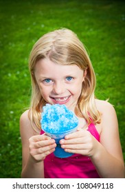 A Young Caucasian Blond Girl Holds A Blue Snow Cone As She Smiles At The Camera.