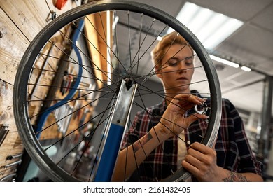 Young caucasian blonde female cycling serviceman checking bicycle wheel spoke with bike spoke key in modern workshop. Bike service, repair and upgrade. Garage interior with tools and equipment