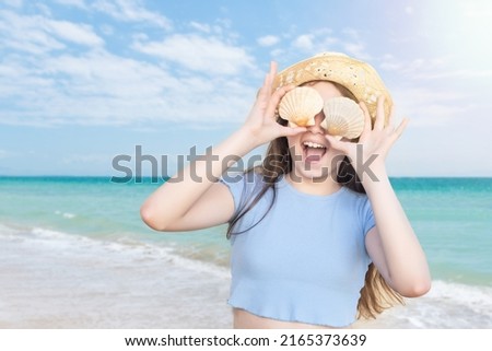 Young Caucasian blonde girl covering her eyes with two shells and smiling with laughter on a turquoise water beach