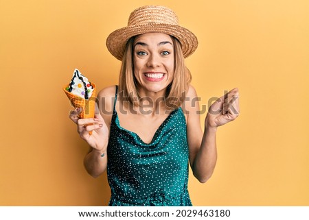 Young caucasian blonde woman eating ice cream screaming proud, celebrating victory and success very excited with raised arm 