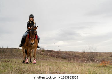 Young Caucasian Blonde Woman Female Girl On The Horse Riding In Nature Wearing Helmet In Winter Or Autumn Day Against A Cloudy Sky In The Field - Freedom Concept Copy Space