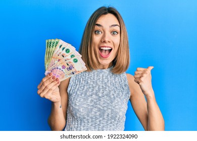Young caucasian blonde woman holding argentine pesos banknotes pointing thumb up to the side smiling happy with open mouth 