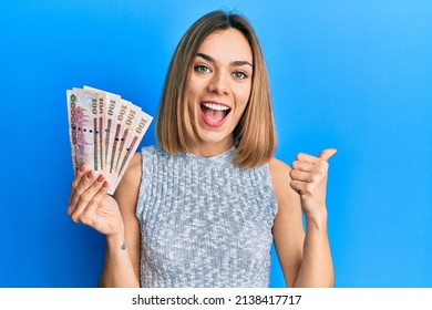 Young Caucasian Blonde Woman Holding Saudi Arabia Riyal Banknotes Pointing Thumb Up To The Side Smiling Happy With Open Mouth 