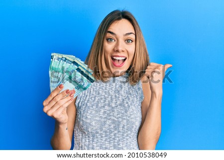 Young caucasian blonde woman holding 50 malaysia ringgit banknotes pointing thumb up to the side smiling happy with open mouth 