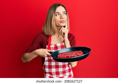 Young Caucasian Blonde Woman Wearing Cook Apron Cooking Meat On Pan Serious Face Thinking About Question With Hand On Chin, Thoughtful About Confusing Idea 