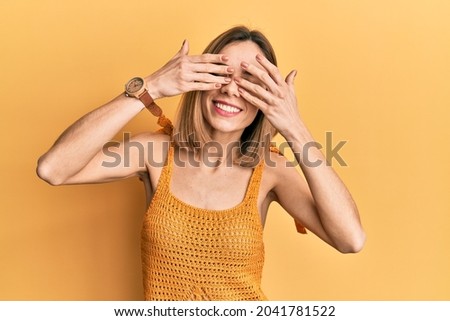 Young caucasian blonde woman wearing casual yellow t shirt covering eyes with hands smiling cheerful and funny. blind concept. 