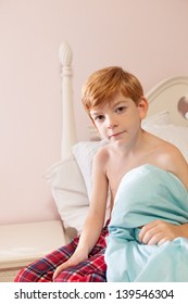 Young Caucasian Boy With Red Hair Sitting On Edge Of Bed In Pajama Bottoms Looking At Camera.