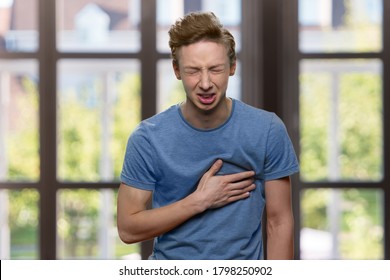 Young Caucasian Boy Suffering From Sudden Heart Attack. Stressed Teen Guy Having Chest Pain On Windows Background. Health Problem Concept.