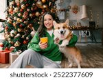 Young caucasian brunette woman looking at the camera sitting with corgi dog by christmas tree at home. High quality photo