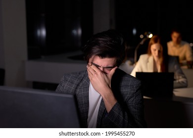 Young Caucasian Brunnete Man In Suit Sits At Night In Office, Eyes Hurt From Overexertion, Suffering From Headache. Male Office Worker Does Not Meet The Deadline. Copy Space