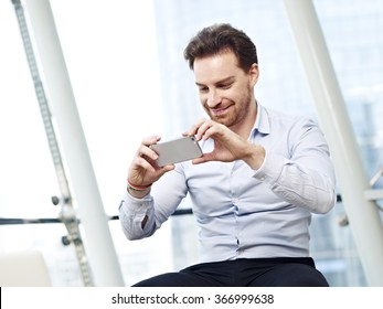 Young Caucasian Business Man Looking At A Picture Or Watching A Video Or Taking A Picture With Mobile Phone And Smiling.