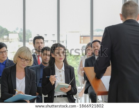 Young caucasian business woman looking to speaker, listening attentively with others and take note while he making a speech in corporate seminar event at conference room.
