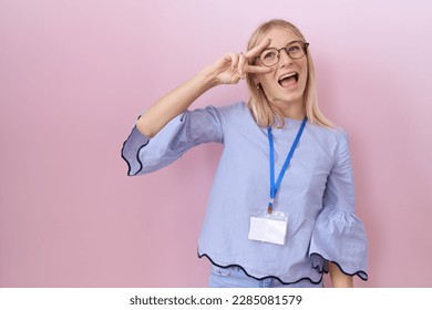 Young caucasian business woman wearing id card doing peace symbol with fingers over face, smiling cheerful showing victory 