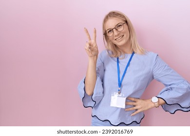 Young caucasian business woman wearing id card smiling looking to the camera showing fingers doing victory sign. number two. 