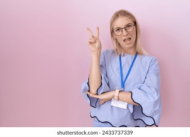 Young caucasian business woman wearing id card smiling with happy face winking at the camera doing victory sign with fingers. number two. 