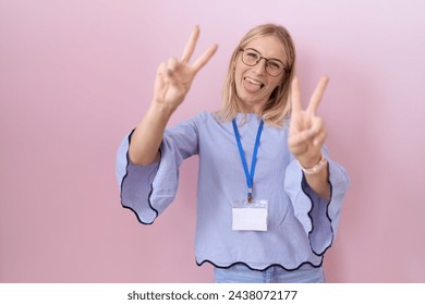 Young caucasian business woman wearing id card smiling with tongue out showing fingers of both hands doing victory sign. number two. 