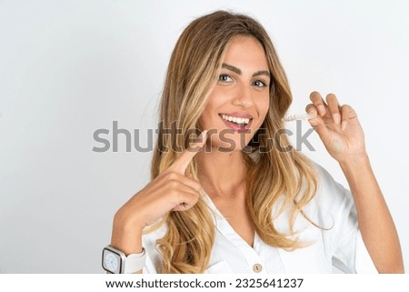 Young caucasian business woman wearing white shirt over white background holding an invisible aligner and pointing to her perfect straight teeth. Dental healthcare and confidence concept.