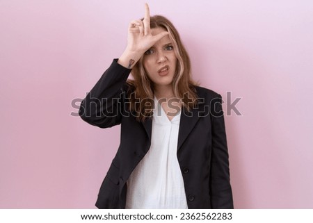 Young caucasian business woman wearing black jacket making fun of people with fingers on forehead doing loser gesture mocking and insulting. 