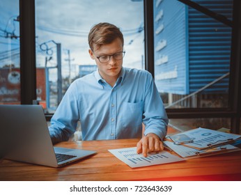 Young Caucasian businessman in denim shirt is standing in office at table and is using laptop with charts, graphs and diagrams on screen. On table is smartphone and charts. Man working.  - Shutterstock ID 723643639