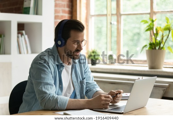 Young Caucasian businessman in earphones work\
on computer online talk speak on video call with client. Male\
employee in headphones use laptop have webcam digital conference\
with customer.