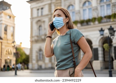 A young Caucasian businesswoman wearing a face mask is taking a walk to work and having a conversation over the phone. - Shutterstock ID 2394492229