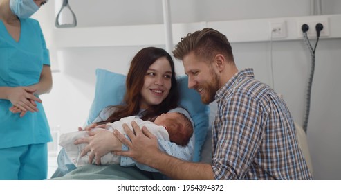 Young Caucasian Couple With Doctor Holding Newborn Baby In Hospital Ward. Nurse Wearing Protective Mask Giving Infant To Happy Parents In Clinic Room. Healthcare Concept