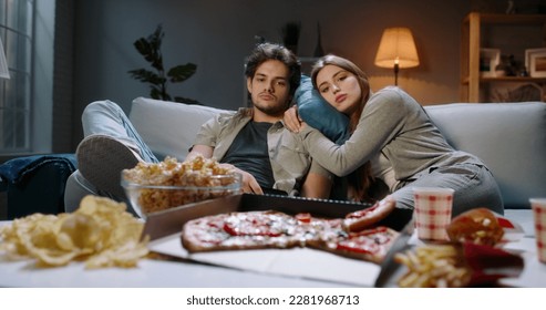 Young caucasian couple extremely frustrated. Bored students are sitting in front of tv, switching channels with remote and eating unhealthy food, tired after hard day  - Powered by Shutterstock