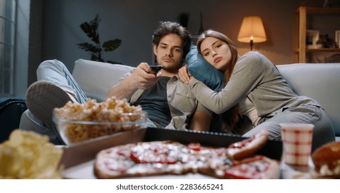 Young caucasian couple extremely frustrated. Bored students are sitting in front of tv, switching channels with remote and eating unhealthy food, tired after hard day  - Powered by Shutterstock