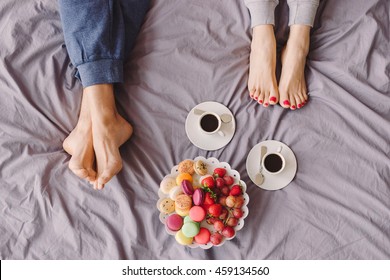 Young Caucasian Couple Having Romantic Breakfast In Bed. Closeup Of Female And Male Feet, Two Cups Of Coffee, Fruits And Colorful Biscuits.