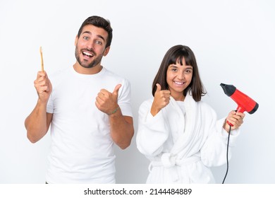 Young Caucasian Couple Holding A Hairdryer And Toothbrush Isolated On White Background Giving A Thumbs Up Gesture With Both Hands And Smiling