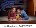 Young caucasian couple lying down on the living room floor using phone and looking at it at nighttime, having fun watching social media together at home.