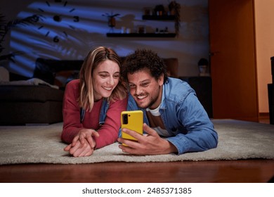 Young caucasian couple lying down on the living room floor using phone and looking at it at nighttime, having fun watching social media together at home. - Powered by Shutterstock