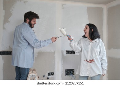 Young Caucasian Couple Playing Sword Fighting With Paint Rollers While Painting Walls In Their New House In Construction.