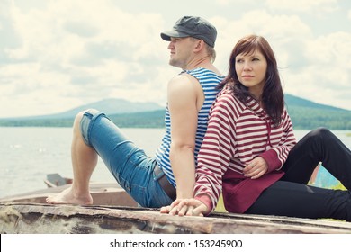 Young Caucasian Couple Sitting Back To Back On An Old Fishing Boat On A Lake Shore