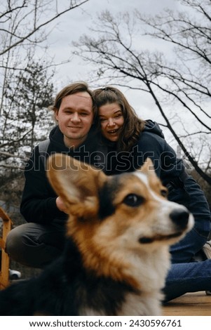 A young Caucasian couple sitting on a wooden bridge in the park, smiling at each other with their dog welsh corgi pembroke. They look happy and relaxed in casual clothes. The dog's face in close-up