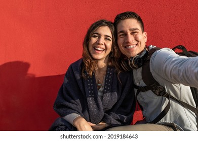 Young caucasian couple take a selfie photo laughing. Man and woman with casual clothing, travelers or students. Red wall on background.