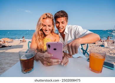 Young caucasian couple using a smart phone to browse on internet sitting at a beach bar during their summer vacation on the coast. Boyfriend and girlfriend booking a hotel room online with a cellphone - Powered by Shutterstock