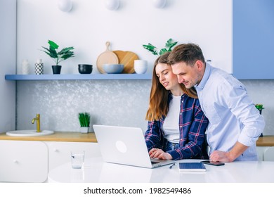 Young Caucasian Couple Of Woman And Man Sitting At Home Kitchen Looking To Computer Laptop Screen Smiling Laughing Pointing To Screen Excited Cheerful Happy. Couple Buying Products Or Services Online